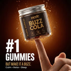 A hand holds a small clear jar of Hebe Brand 'Buzz Cola' Vijaya Gummies against a dark brown background with floating droplets. The jar has a black label with gold lettering and a silver metal lid.